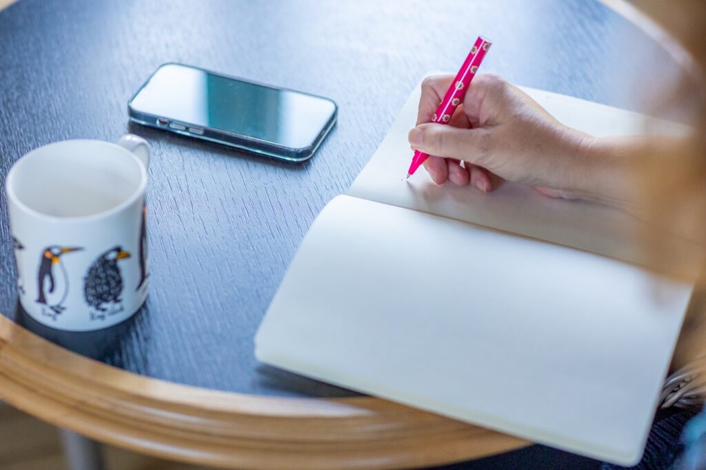notebook, mug and phone, with person writing in notebook with a pink pen